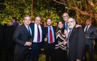 Um grupo de seis pessoas está reunido em um evento formal, ao ar livre, com plantas verdes e iluminação quente ao fundo. Cinco homens e uma mulher estão posando para a foto, todos vestidos com trajes sociais.