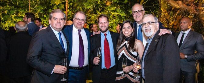 Um grupo de seis pessoas está reunido em um evento formal, ao ar livre, com plantas verdes e iluminação quente ao fundo. Cinco homens e uma mulher estão posando para a foto, todos vestidos com trajes sociais.