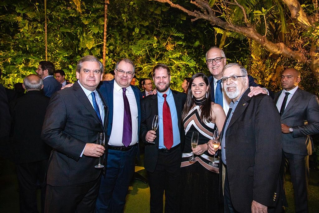 Um grupo de seis pessoas está reunido em um evento formal, ao ar livre, com plantas verdes e iluminação quente ao fundo. Cinco homens e uma mulher estão posando para a foto, todos vestidos com trajes sociais.